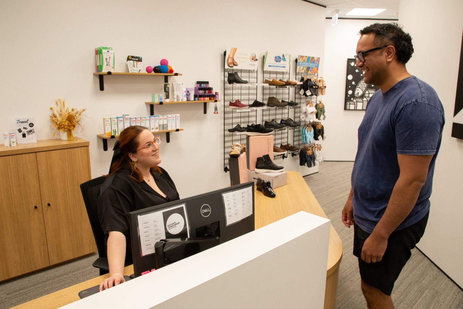 A patient chatting with a friendly Capital Podiatry receptionist at the front desk, with the footwear display behind