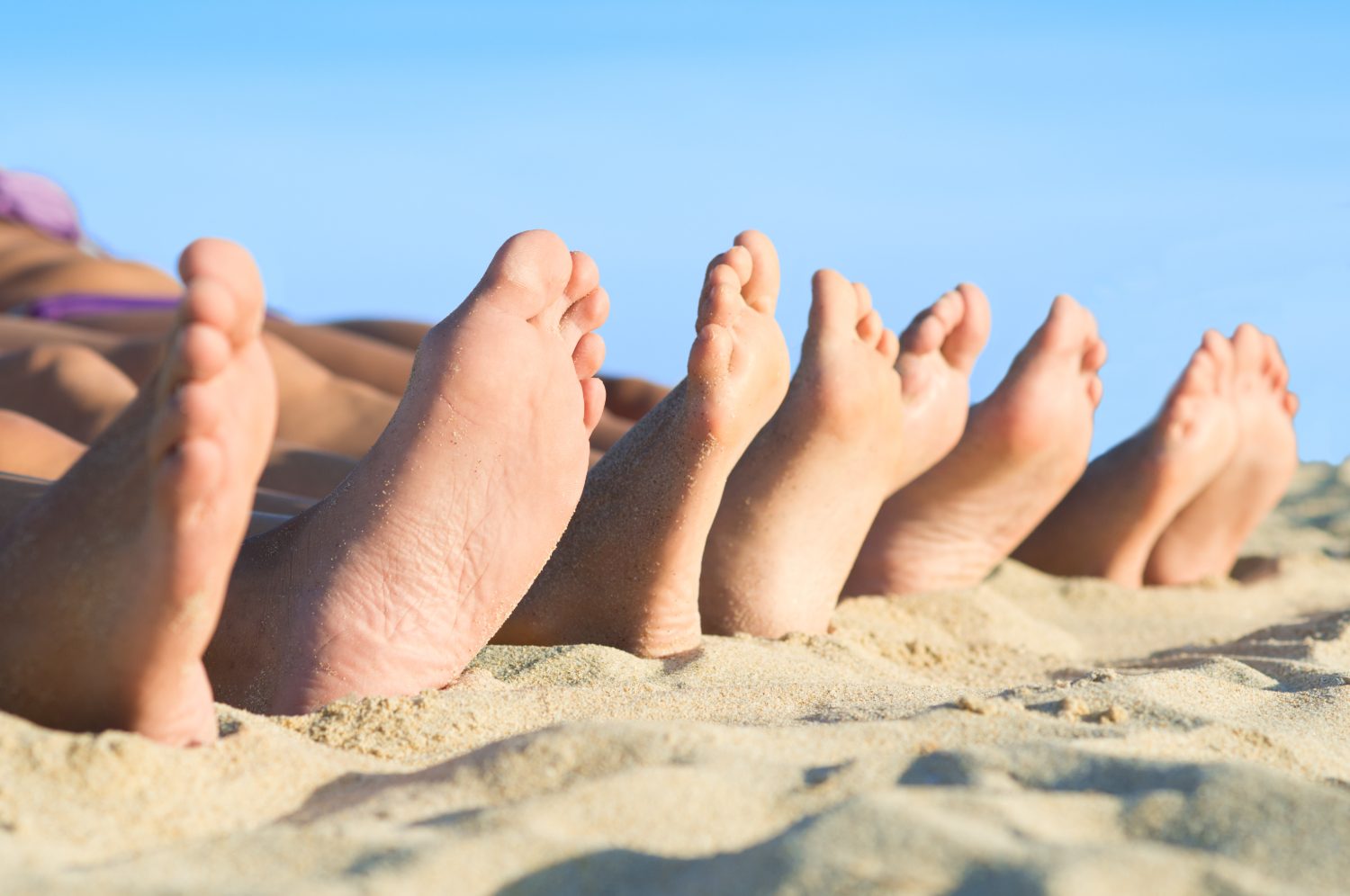 A row of relaxed bare feet resting in the sand under a clear blue sky
